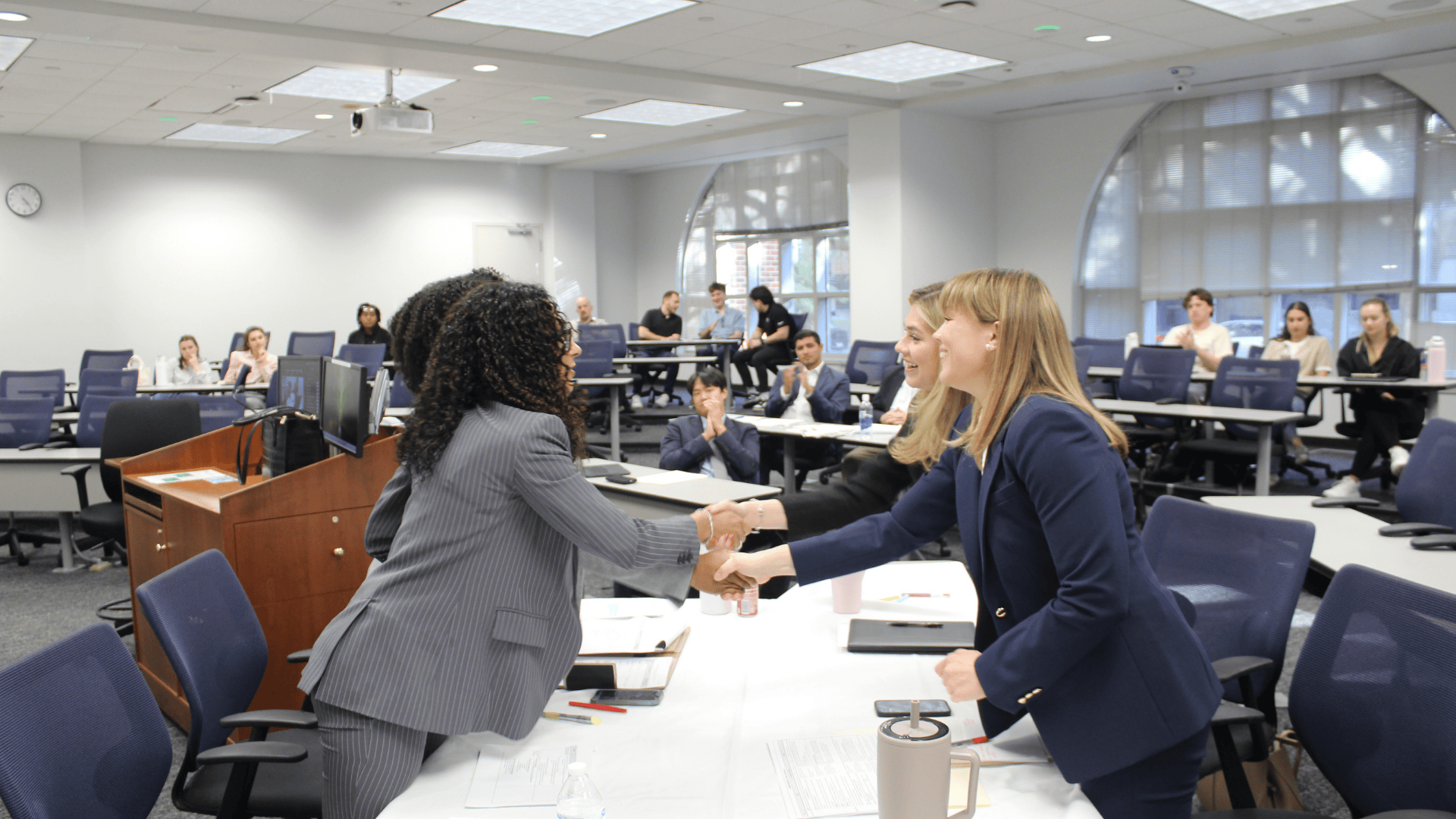 Four competitors shake hands, smiling, in a meeting room.
