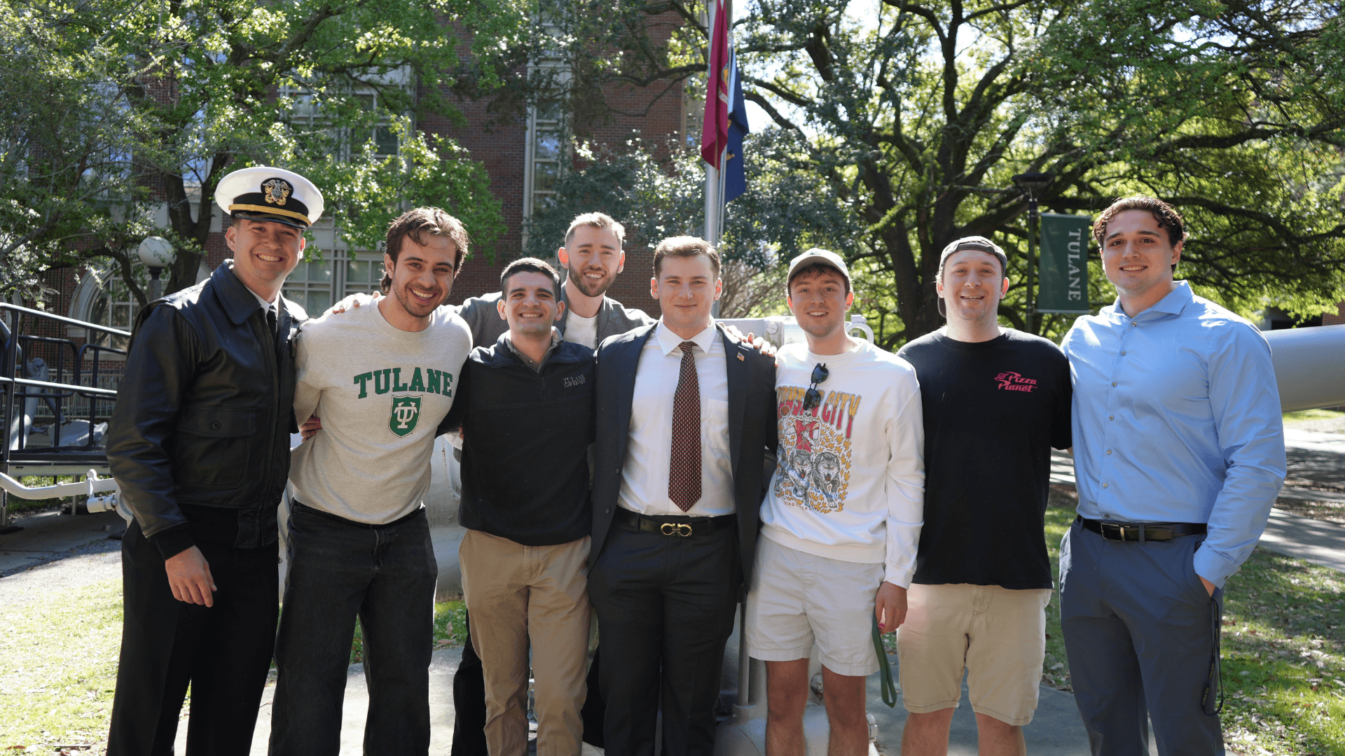 A group of students, one in a naval uniform, others in casual clothes, smiling outdoors.