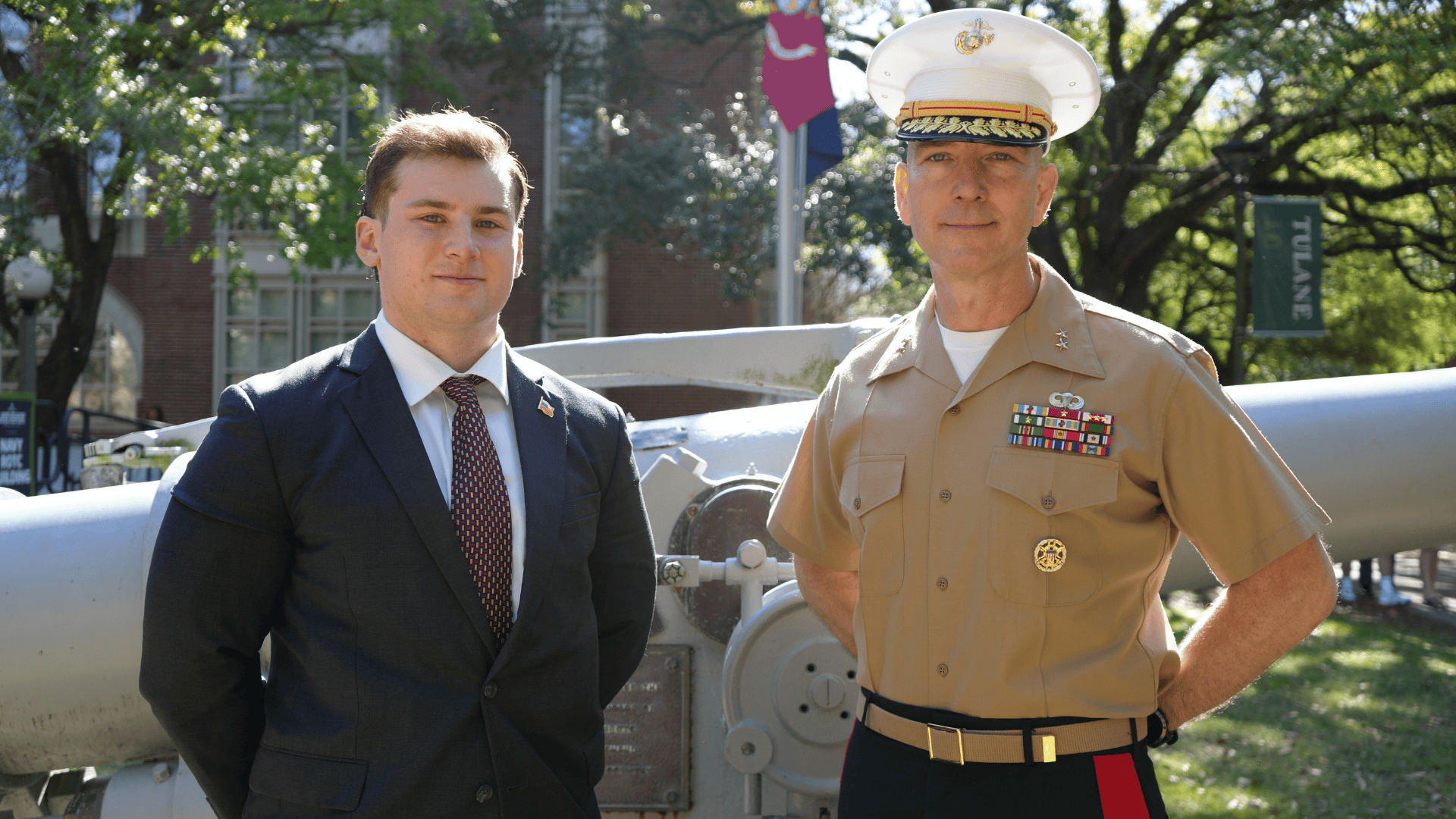 Man in suit and Marine officer stand together outdoors with a cannon.