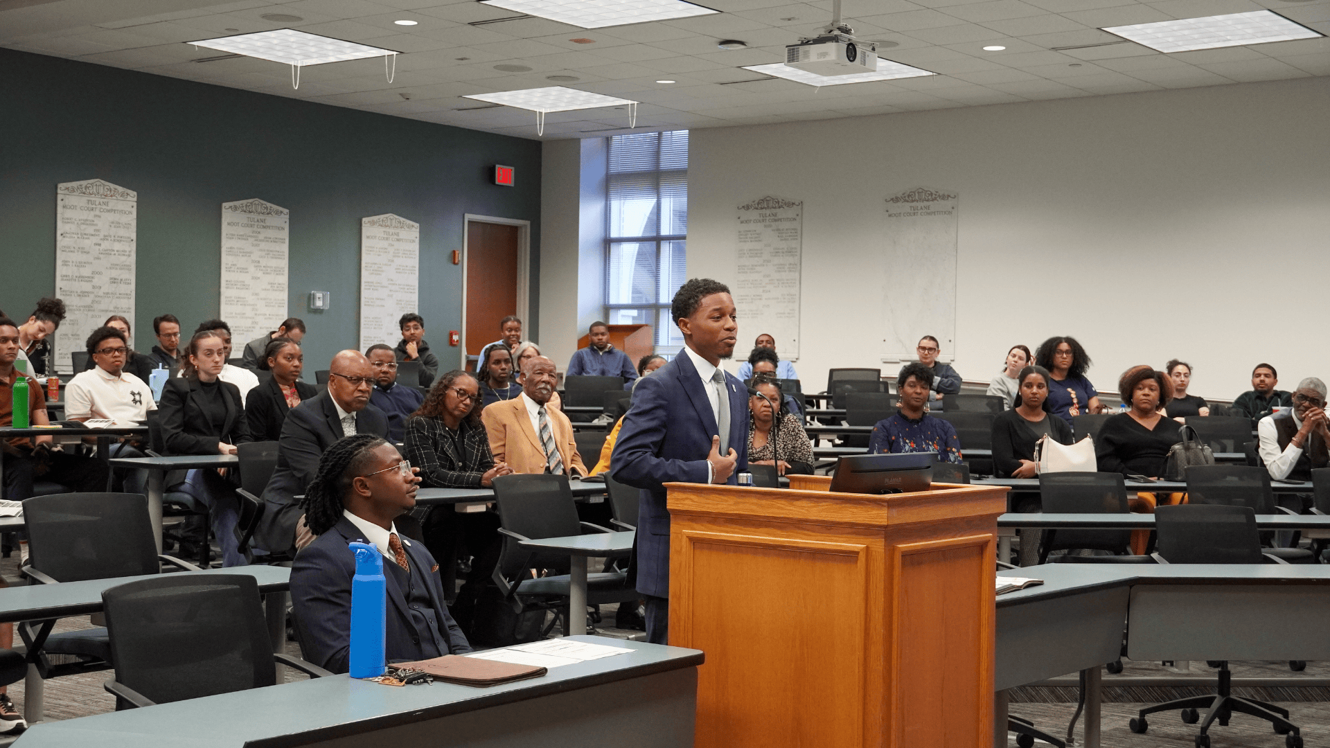 Man at podium addresses seated audience in a lecture or courtroom setting.
