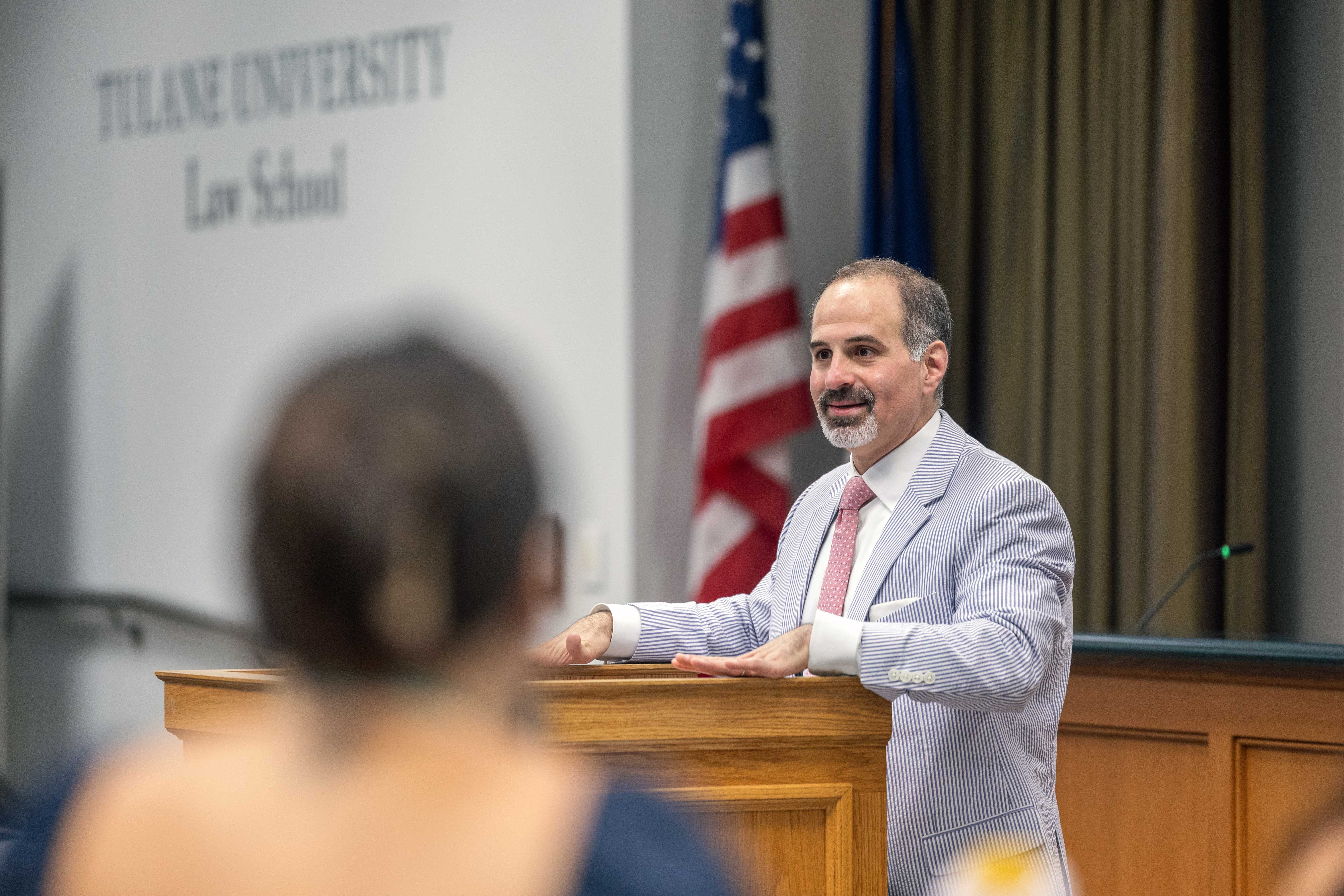 Man in striped jacket speaks from podium; Tulane Law School banner and US flag in background.