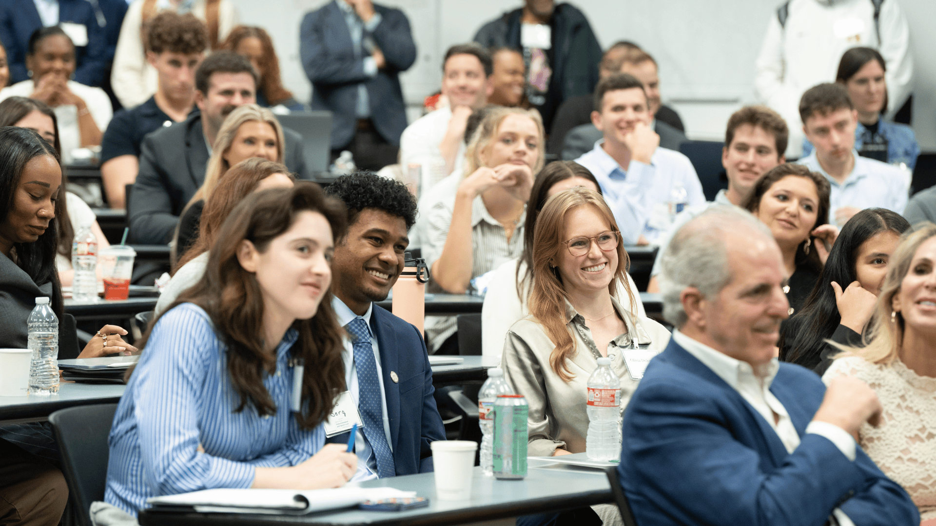 A diverse group of smiling students and adults attentively watching a presentation in a lecture hall.