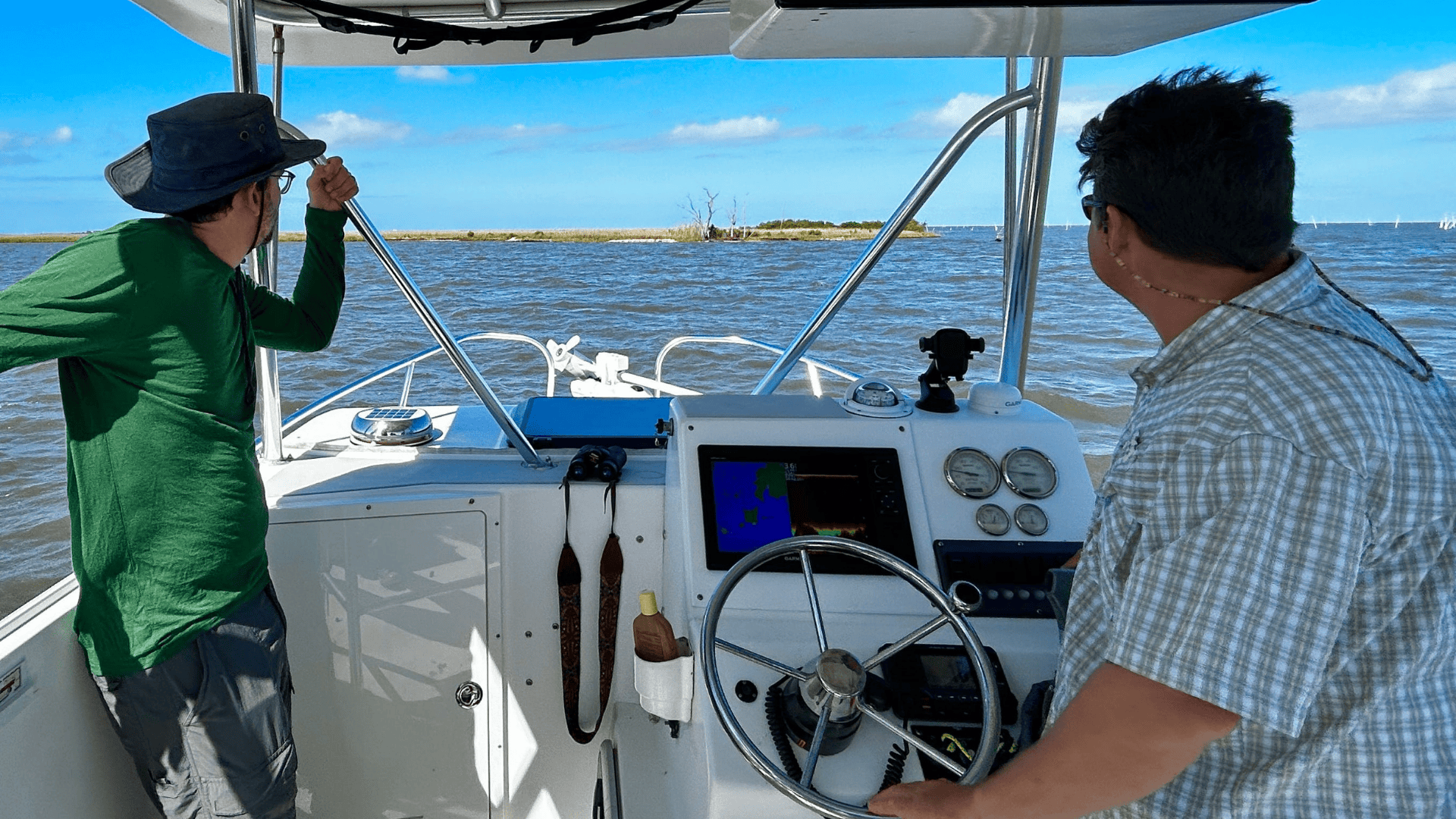 Two men on a white boat, one steering, the other looking at the distant shore.