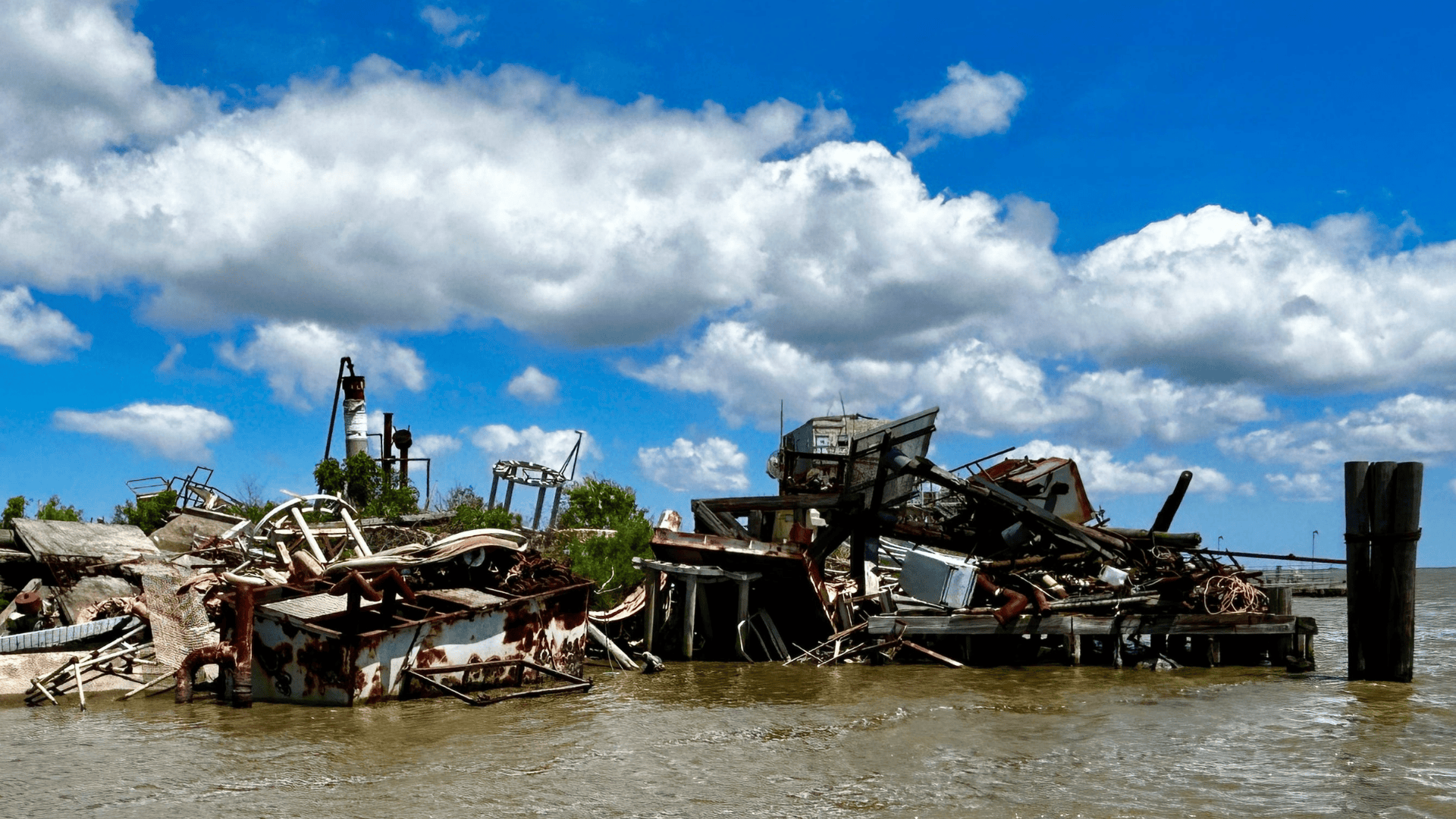 Dilapidated, rusted structures and debris in murky water under a blue, cloudy sky.