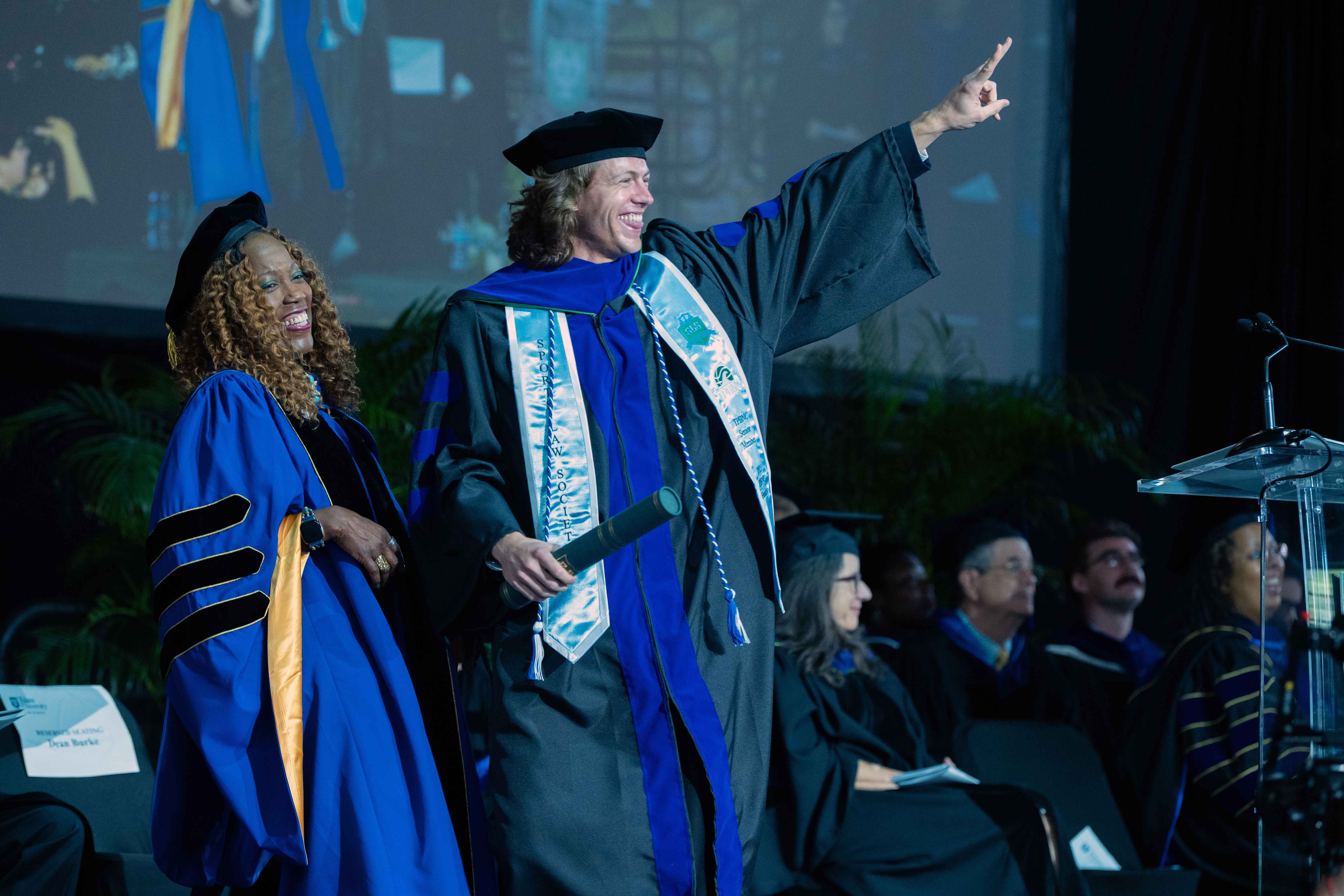 Photograph: Man in academic regalia points excitedly, smiling woman beside him at graduation.