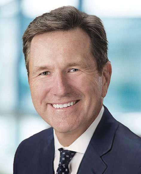Smiling middle-aged man in a dark blue suit and patterned tie with a blurred office background.
