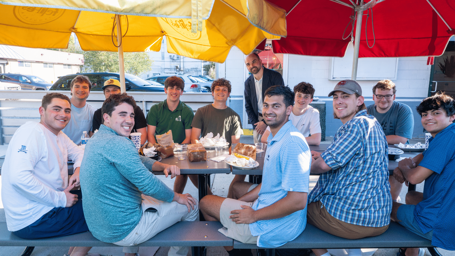 Smiling young men sit at an outdoor restaurant table under colorful yellow and red umbrellas.