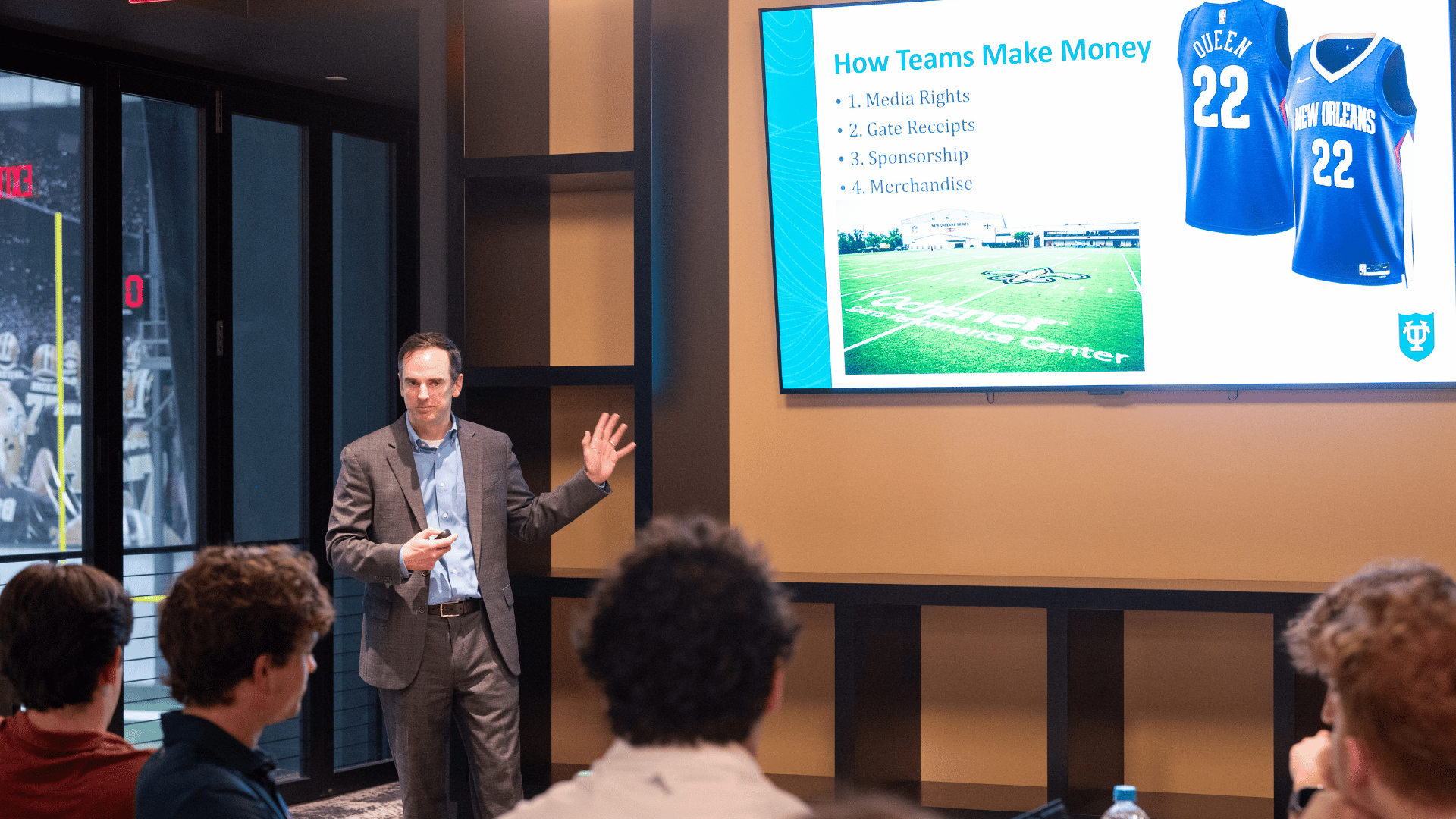 Man in a suit presents to students, gesturing at a screen displaying sports jerseys and a field graphic.