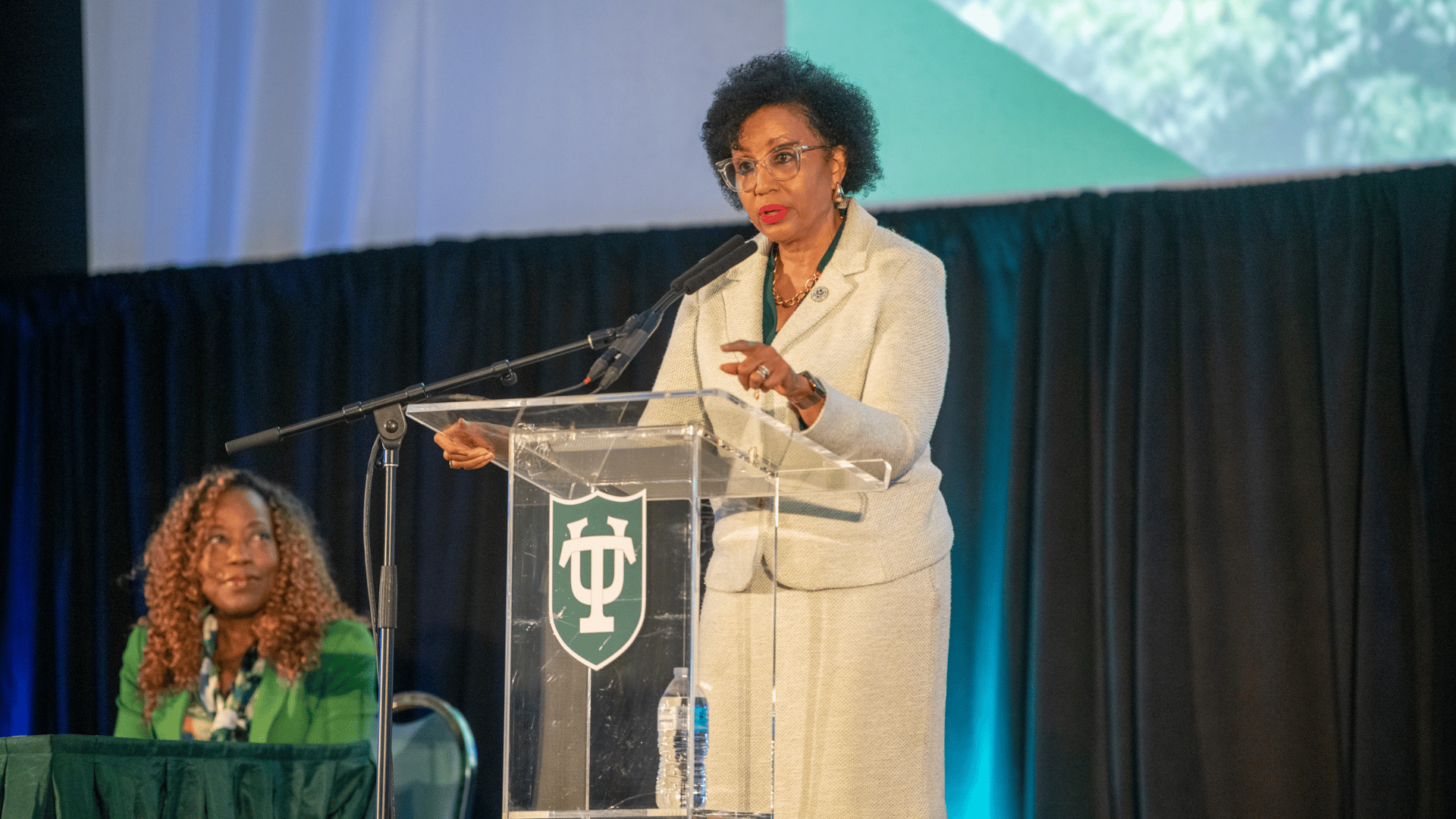 Woman in light suit speaking at a podium with a green logo; another woman seated.