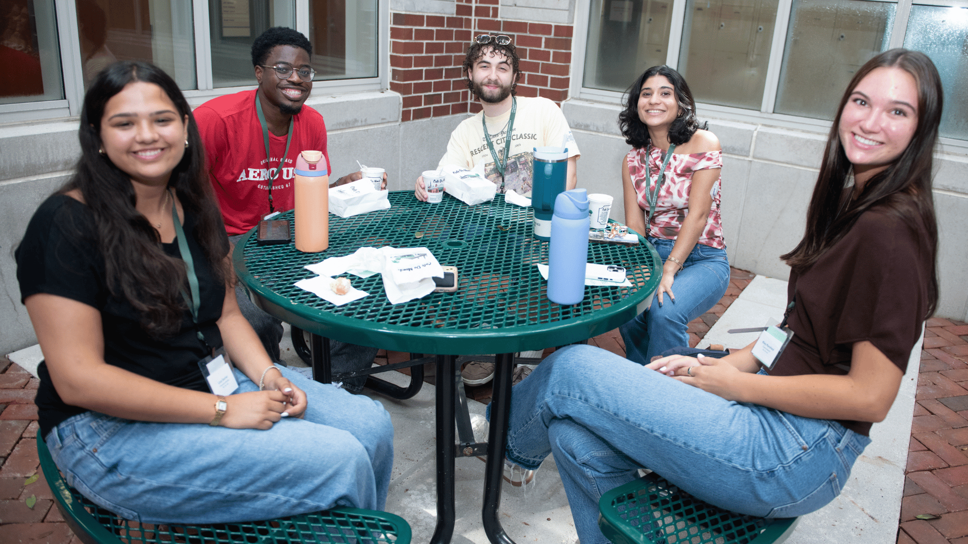 Five smiling young adults sit around a green outdoor table in a courtyard.