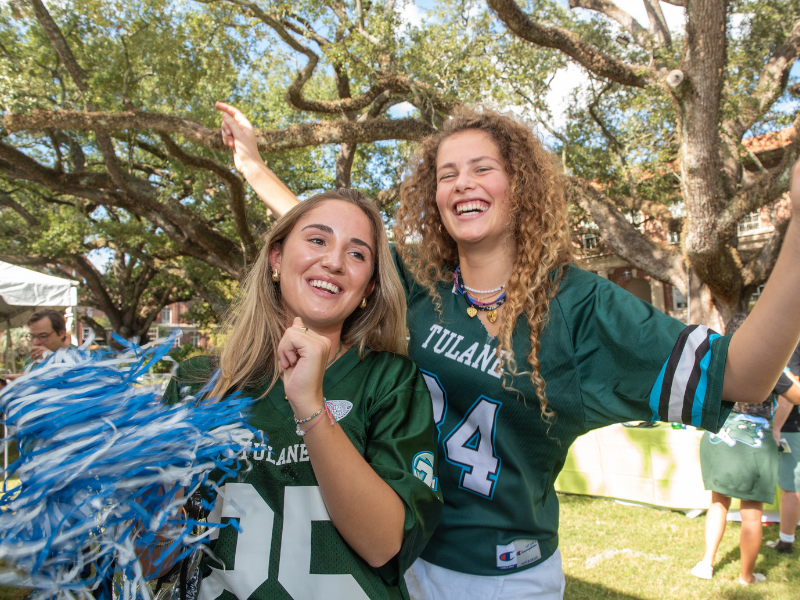 Two cheerful women in green Tulane jerseys, one holding blue pom-poms.