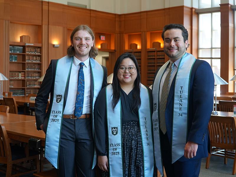 Jack Dean, Katie Chong, and Pablo Gonzales pose for a photo in the Law Library. They wear blue and green stoles that read "Tulane 34" and "Class of 2025"
