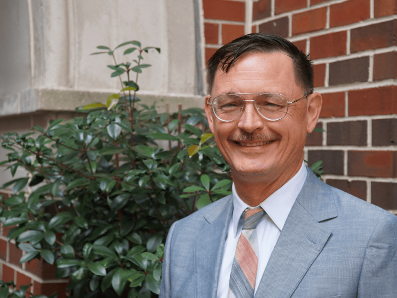 Professor Brian Frye poses for a headshot against a red brick wall and green foliage.