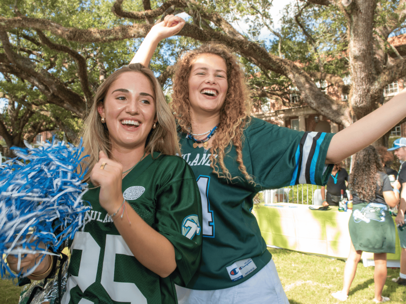 Tulanians smile and cheer while wearing football jerseys and holding pom poms at the tailgate.