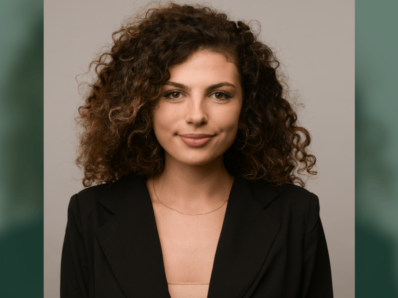 Professional photograph of a woman with curly brown hair, subtle smile, wearing a black blazer.