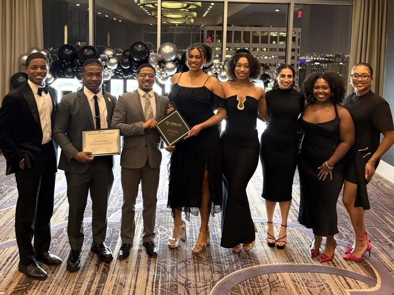 Eight formally dressed people, two holding awards, pose at an elegant event.