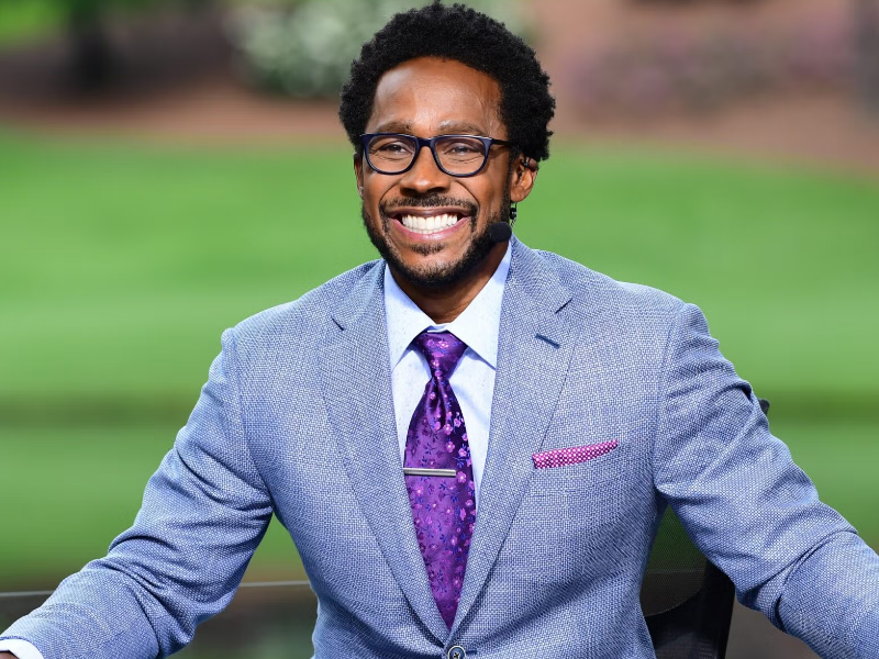 Desmond Howard in light blue suit, purple tie, and glasses, golf course background.