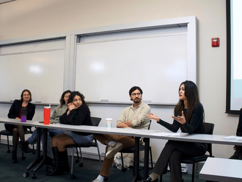 Five panelists sit at a table in a classroom, one woman actively speaks.