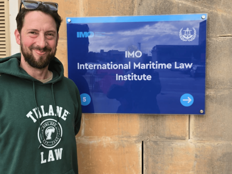 Man in green Tulane Law hoodie smiles next to the IMO International Maritime Law Institute sign.