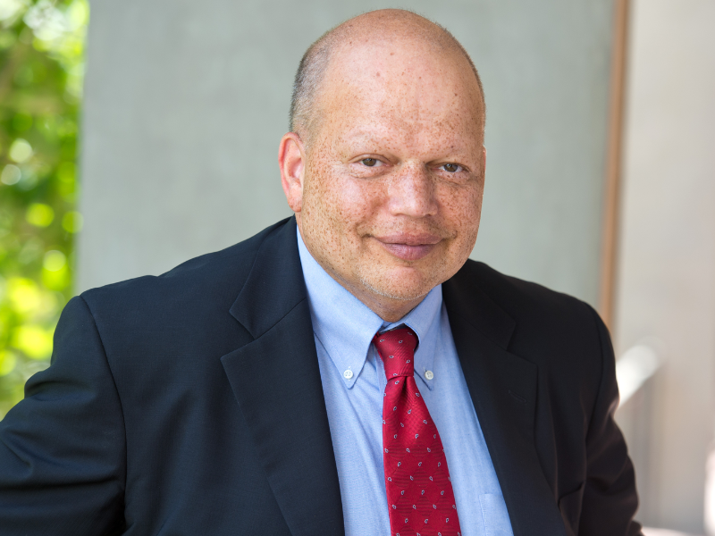 Smiling bald man in a black suit, blue shirt, and red tie.