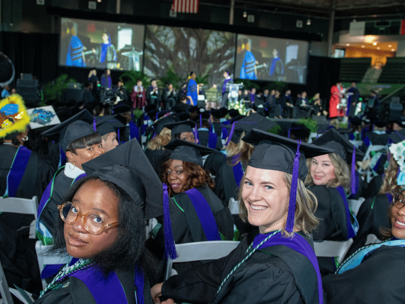 Photograph: Smiling graduates in black, purple, green gowns at a large ceremony.