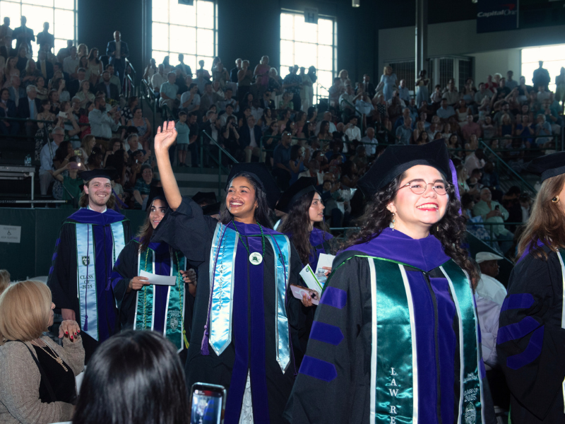 Tulane Law School graduates process into Devlin Fieldhouse wearing graduation gowns and surrounded by a crowd of onlookers in the stands.