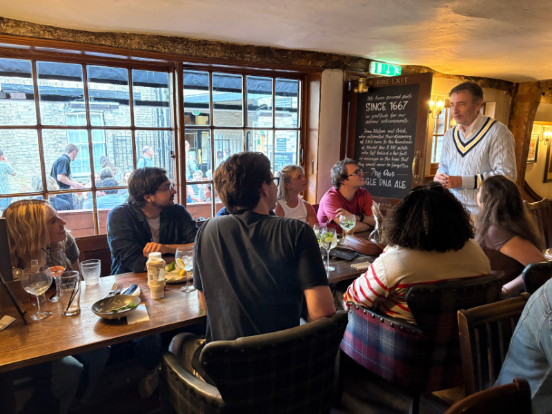 Students sit at a table in a pub while listening to a presenter.