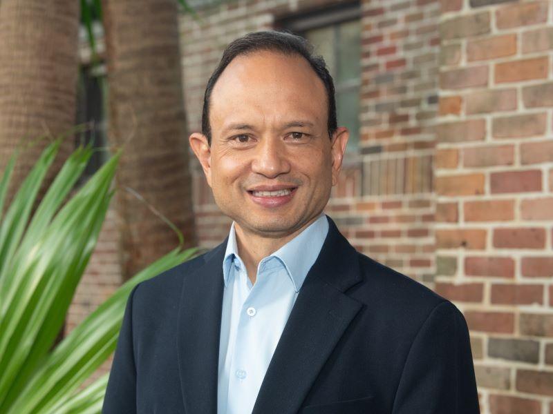 Smiling man in a dark suit and light blue shirt standing outdoors with a brick wall and palm trees in the background.