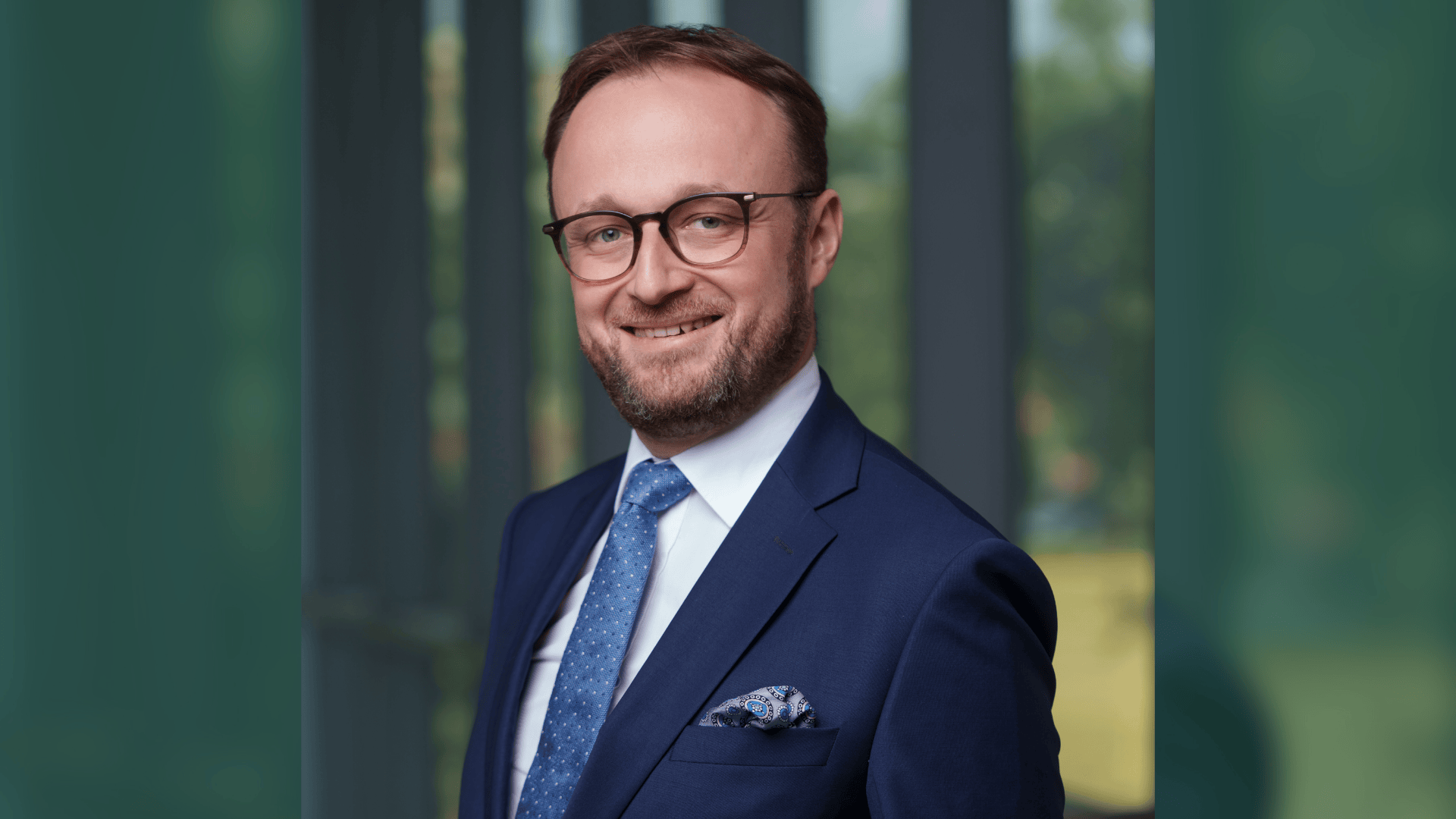 Mateusz in blue suit and glasses against a blurred office background.