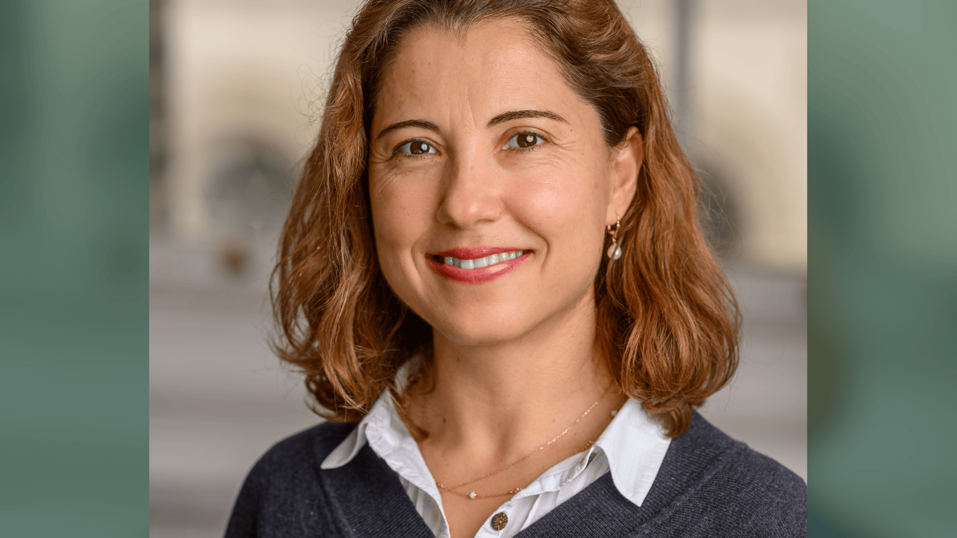 Smiling woman with shoulder-length brown hair, wearing a white shirt, dark sweater, and pearl earrings.