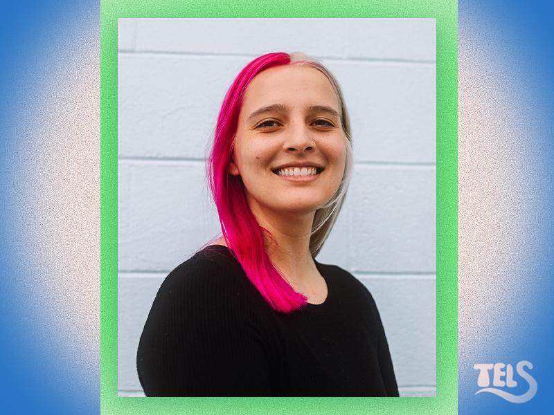 Smiling woman with split pink and blonde hair, wearing a black top, against a light wall.