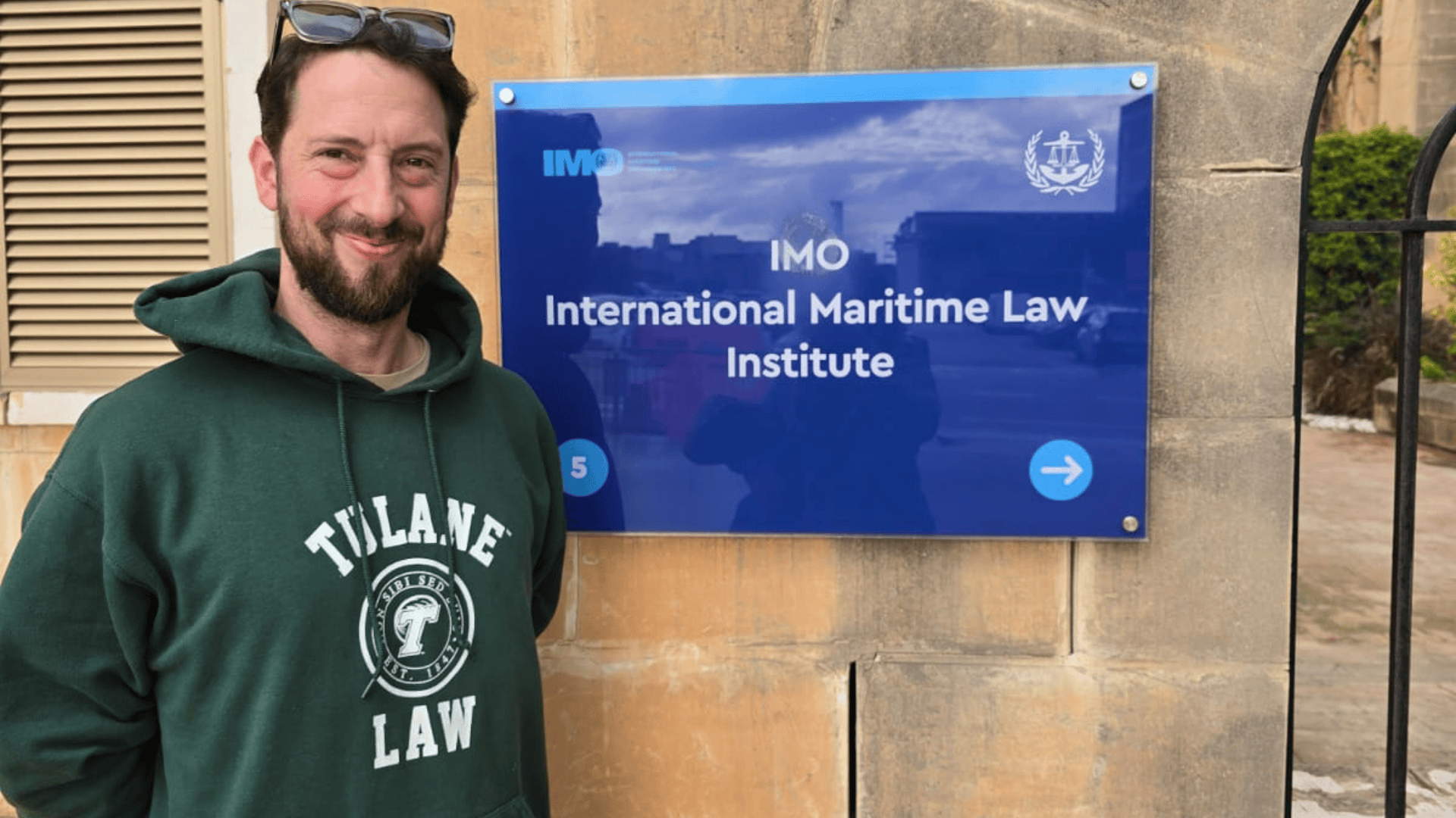 Man in green Tulane Law hoodie smiles next to the IMO International Maritime Law Institute sign.
