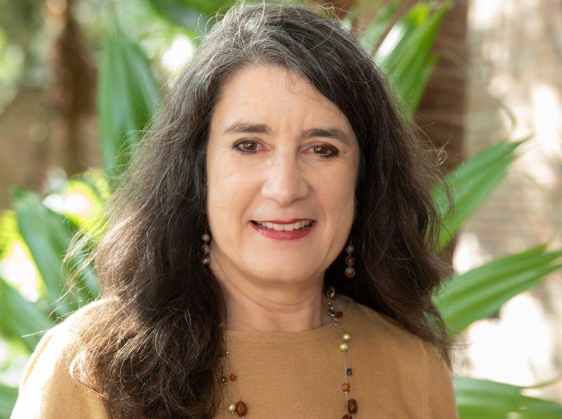 Smiling woman with long dark hair and a brown top, against a green plant background.