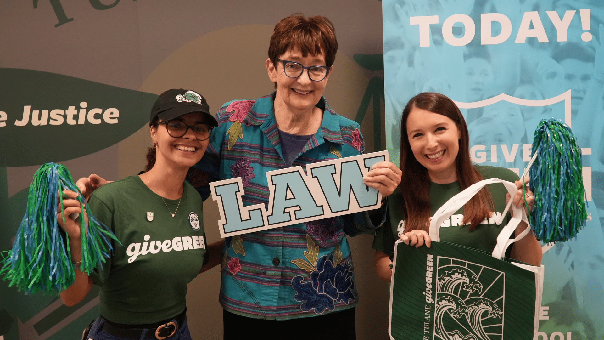 Three women smile, one holding a "LAW" sign, others holding pom-poms and a green tote bag.