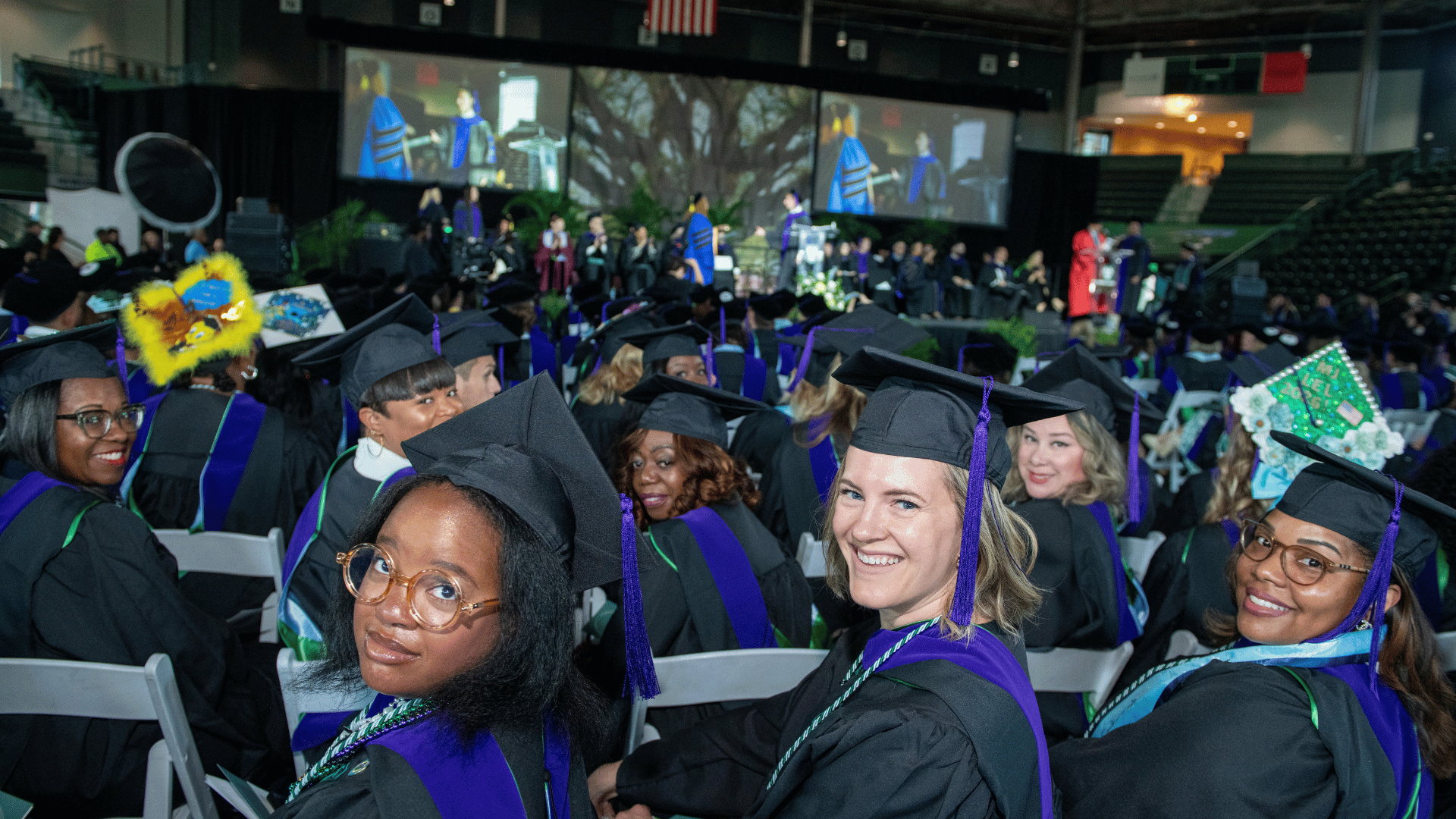 Photograph: Smiling graduates in black, purple, green gowns at a large ceremony.