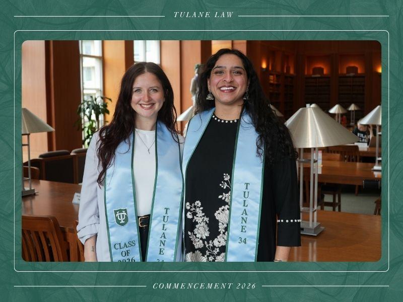 Two smiling graduates in light blue Tulane stoles pose in a library.