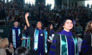 Tulane Law School graduates process into Devlin Fieldhouse wearing graduation gowns and surrounded by a crowd of onlookers in the stands.