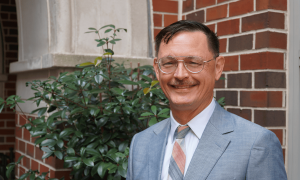 Professor Brian Frye poses for a headshot against a red brick wall and green foliage.