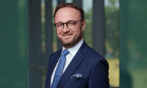 Mateusz in blue suit and glasses against a blurred office background.