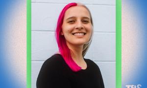 Smiling woman with split pink and blonde hair, wearing a black top, against a light wall.