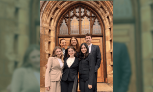 Six young adults in professional attire smile in front of an ornate stone archway.