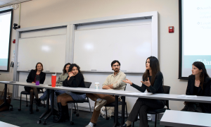 Five panelists sit at a table in a classroom, one woman actively speaks.
