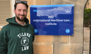 Man in green Tulane Law hoodie smiles next to the IMO International Maritime Law Institute sign.