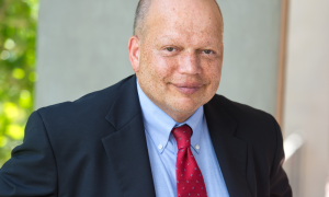 Smiling bald man in a black suit, blue shirt, and red tie.