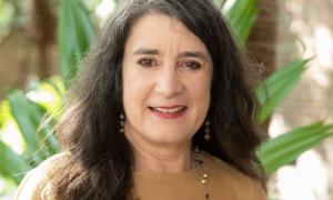 Smiling woman with long dark hair and a brown top, against a green plant background.