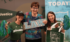 Three women smile, one holding a "LAW" sign, others holding pom-poms and a green tote bag.