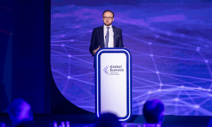 Man in suit speaking at a podium with a "Global Summit" logo on a stage.