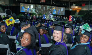 Photograph: Smiling graduates in black, purple, green gowns at a large ceremony.